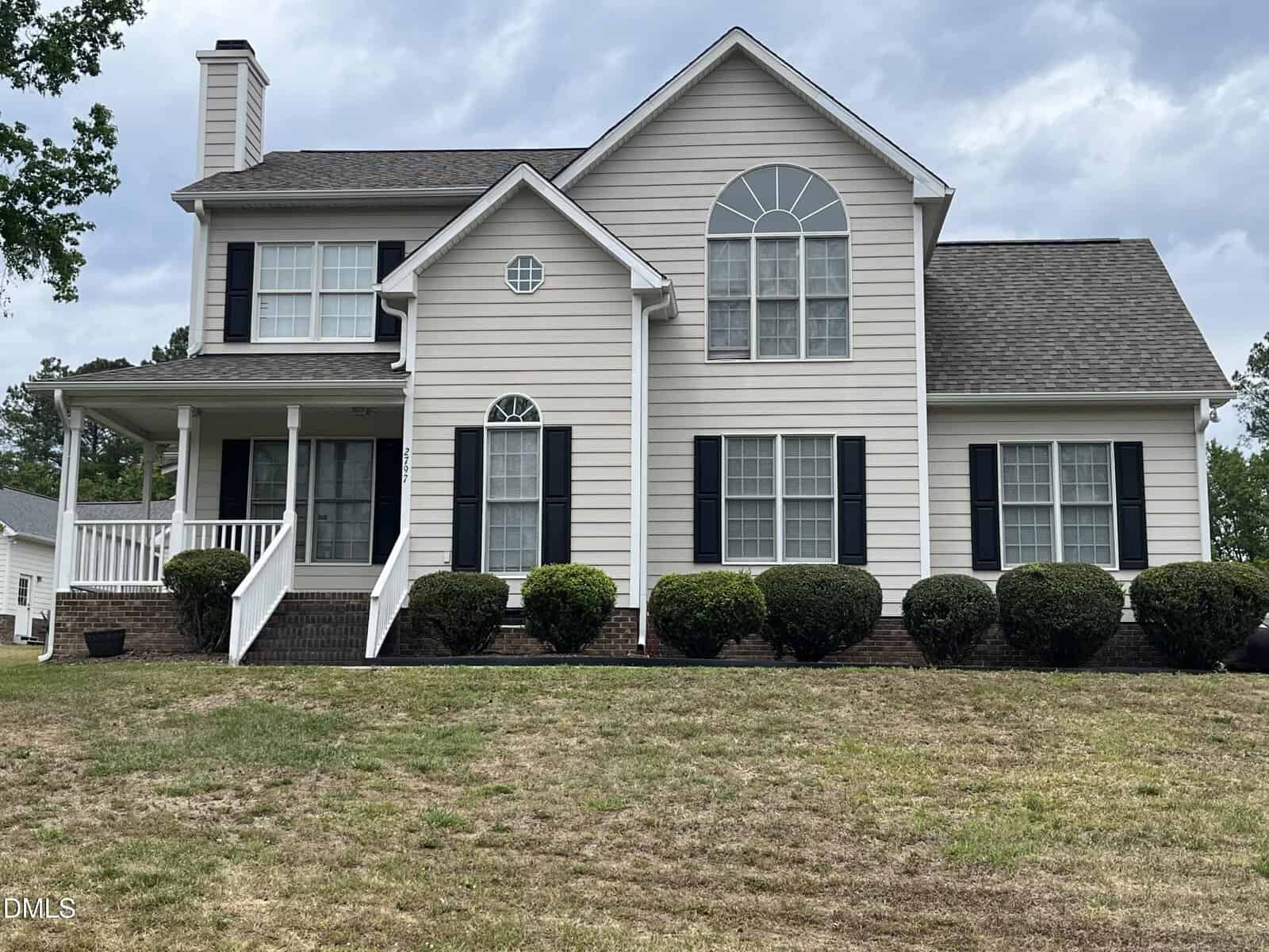 Two-story beige house with dark shutters, arched windows, a front porch with white railings, shrubs along the foundation, and a patchy lawn in front. The sky is cloudy.