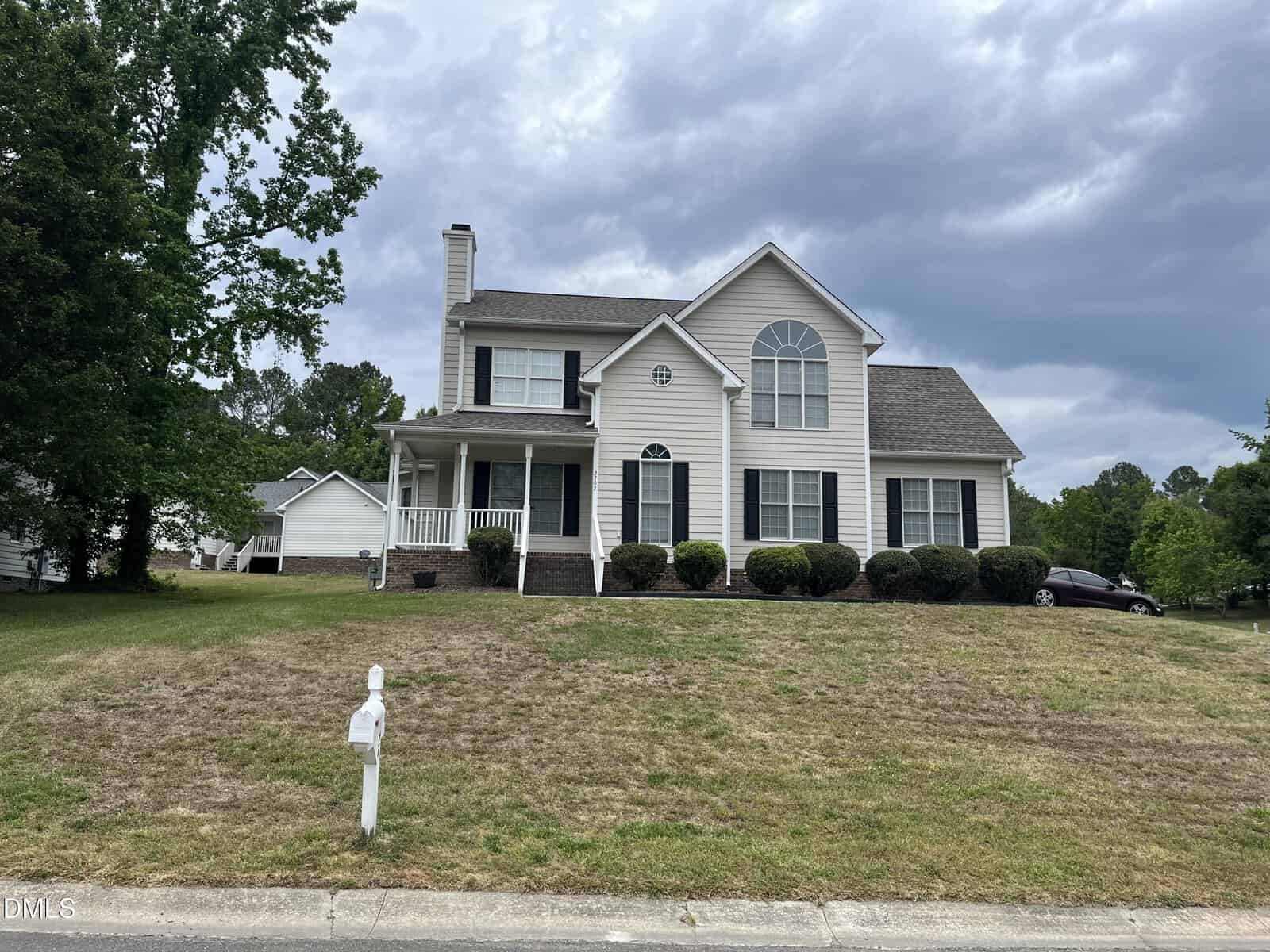 Two-story beige suburban house with white trim, large front windows, porch, and attached garage. Lawn is patchy with brown and green grass. A white mailbox stands near the street. Cloudy sky overhead.