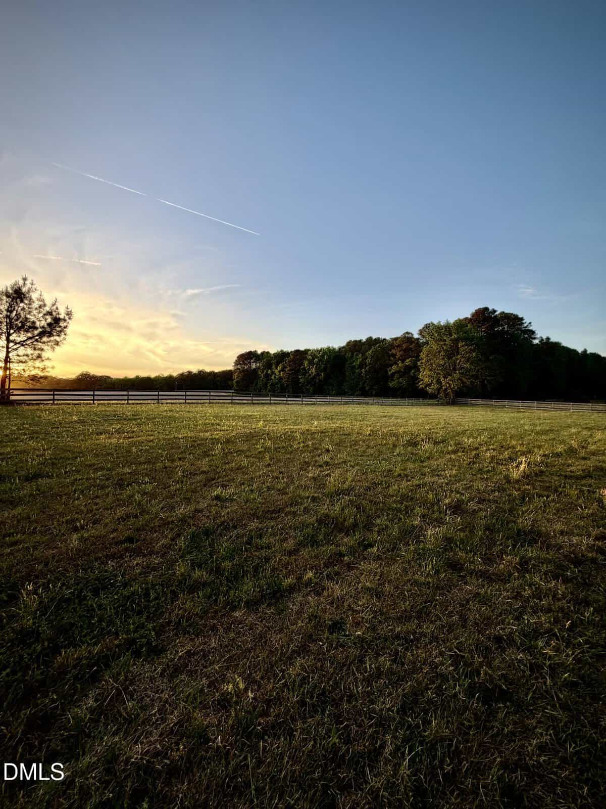 Sunset view of an open grassy field bordered by a wooden fence and trees, with a clear blue sky and a few streaks of clouds overhead. The scene looks peaceful and serene.