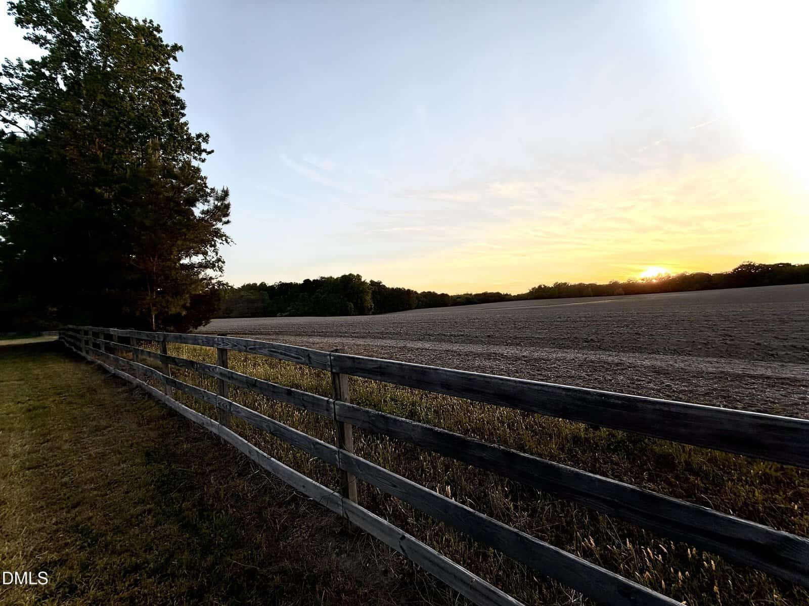 A wooden fence runs alongside a grassy field at sunset, with trees on the left and a clear sky fading from blue to yellow as the sun sets on the horizon.