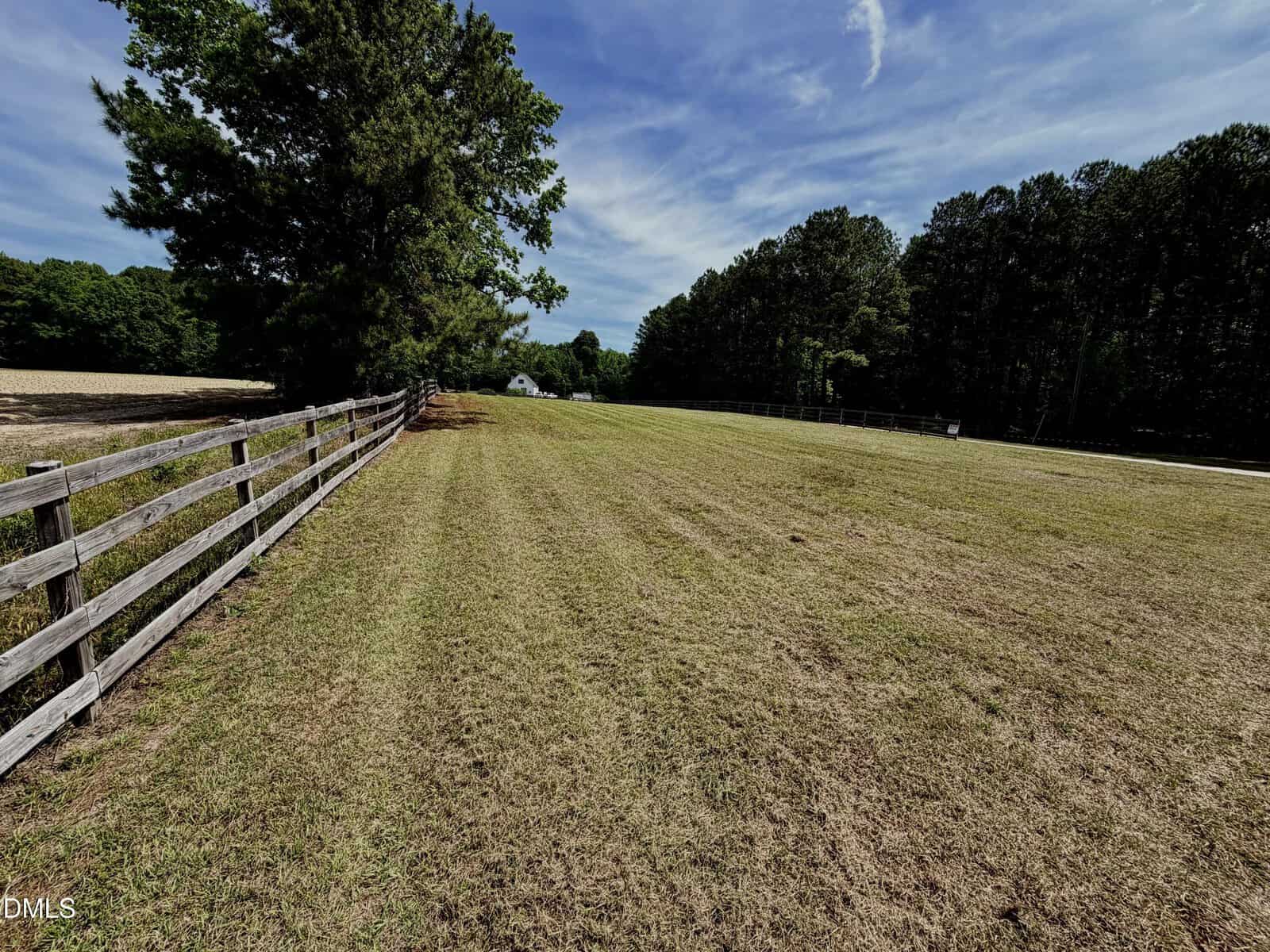 A grassy field with a wooden fence on the left, bordered by trees under a blue sky with wispy clouds; a distant white house is partially visible near the horizon.