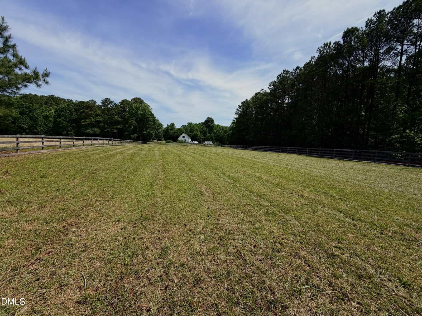 A wide, freshly mowed grassy field is bordered by wooden fences and trees under a blue sky with wispy clouds. A white house or barn is visible in the distance.