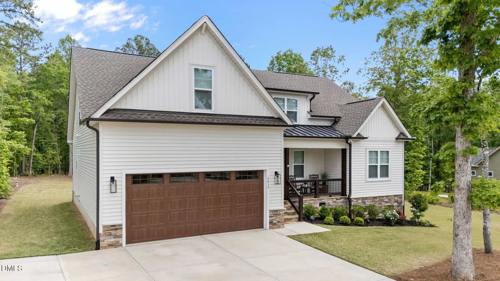 A modern two-story house with white siding, brown garage door, front porch with chairs, and a well-maintained lawn, surrounded by trees and greenery under a blue sky.