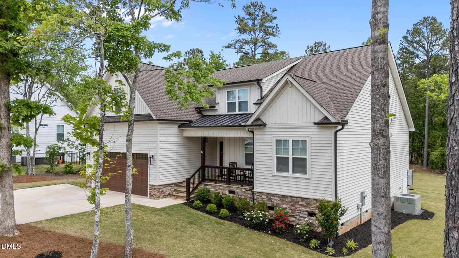 A modern two-story house with light-colored siding, brown shingles, and stone accents. It features a front porch, attached garage, landscaped yard, and is surrounded by trees and greenery.