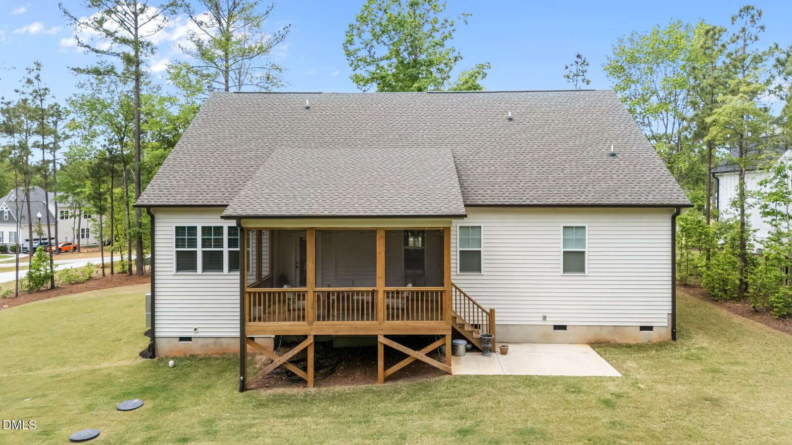 A white house with a gray shingled roof, wooden back porch with stairs and railing, concrete patio, and grassy yard bordered by trees on a sunny day.