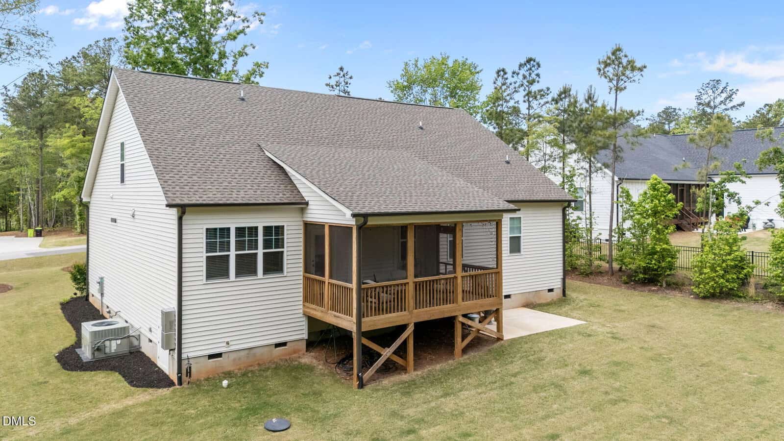 A beige house with a gable roof features a screened-in back porch with wooden railings, set on a grassy yard with trees and neighboring homes in the background under a blue sky.