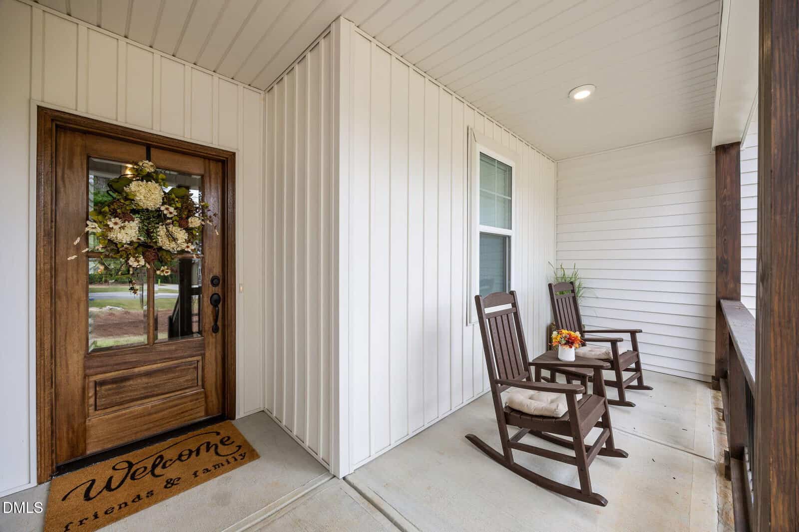 A front porch with two cushioned rocking chairs, a small table with flowers, a wooden door decorated with a floral wreath, and a welcome mat that says, “Welcome friends & family.”.