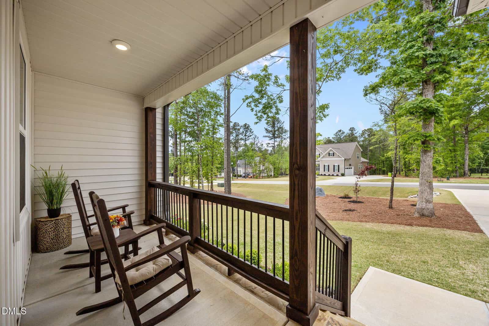 Covered porch with two wooden rocking chairs, a small table with flowers, potted plant, and railing, overlooking a green yard and suburban street with trees and houses in the background.