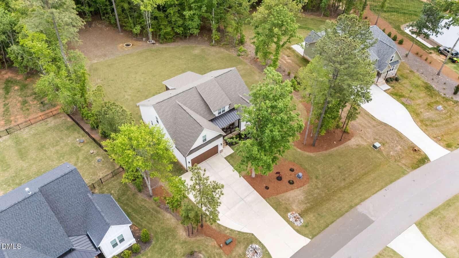 Aerial view of a suburban house with a large backyard, driveway, garage, and surrounding trees. Neighboring homes and lawns are visible, along with a curved street in the foreground.