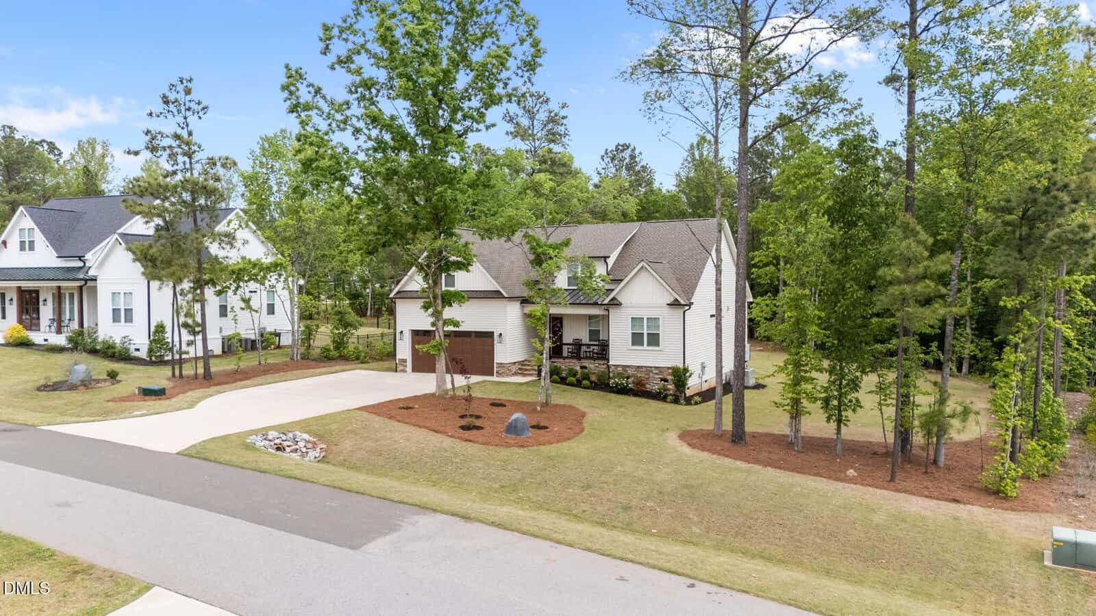 A single-story house with a three-car garage, front porch, and neatly landscaped yard with trees and mulch, situated on a suburban street with neighboring homes and forested area in the background.