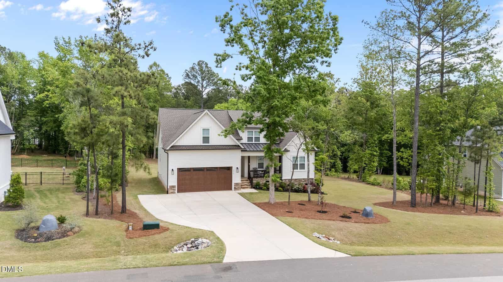 A modern two-story house with a brown double garage, white siding, and a manicured front yard with trees and shrubs, set in a suburban neighborhood beside a wooded area.