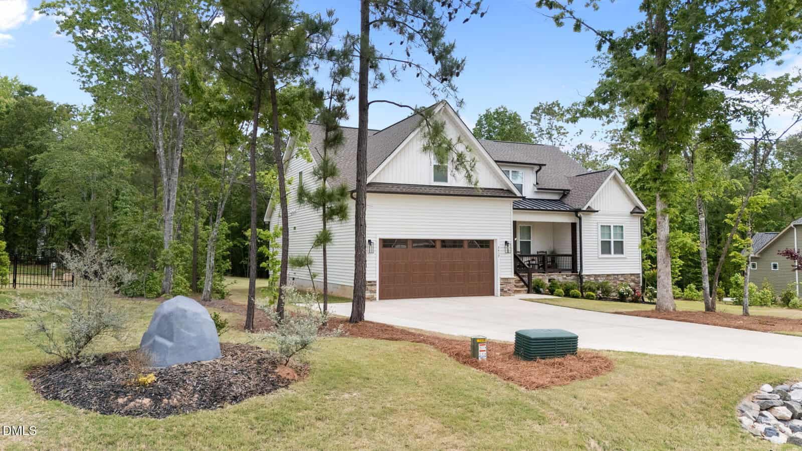 A two-story modern suburban house with a beige exterior, dark brown garage door, and covered front porch, surrounded by trees and landscaped yard with mulch, bushes, and a large gray rock.