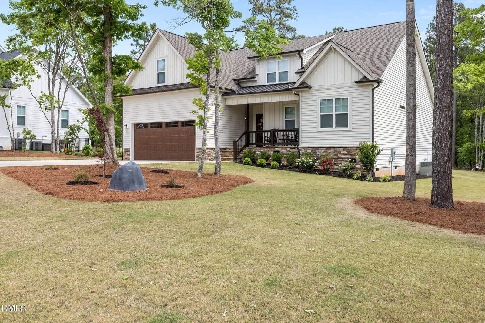 A modern, two-story suburban house with white siding, stone accents, a brown garage door, a covered front porch, and landscaped yard with trees and mulch.