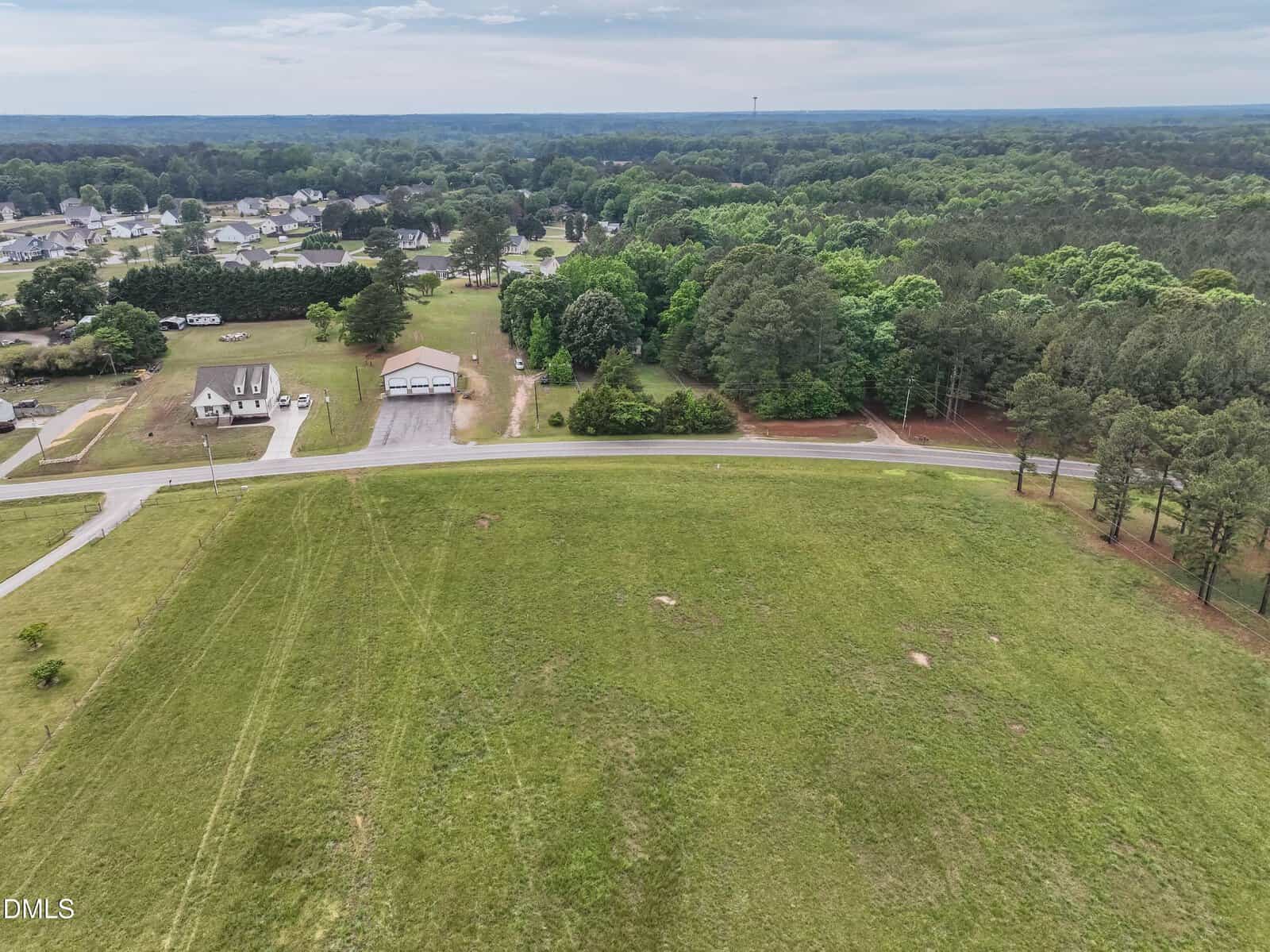 Aerial view of a grassy open field bordered by a road, with houses, trees, and wooded areas in the background under a cloudy sky. Suburban neighborhood visible in the distance.
