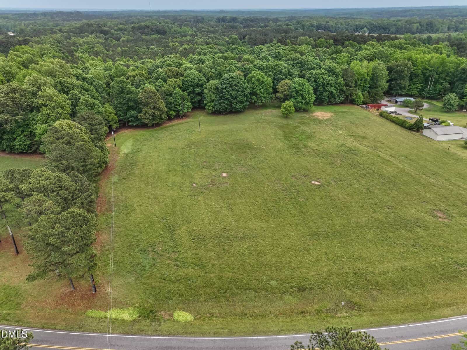Aerial view of a large, grassy open field bordered by dense green trees, with a paved road at the bottom edge and a few scattered structures visible on the right side near the trees.