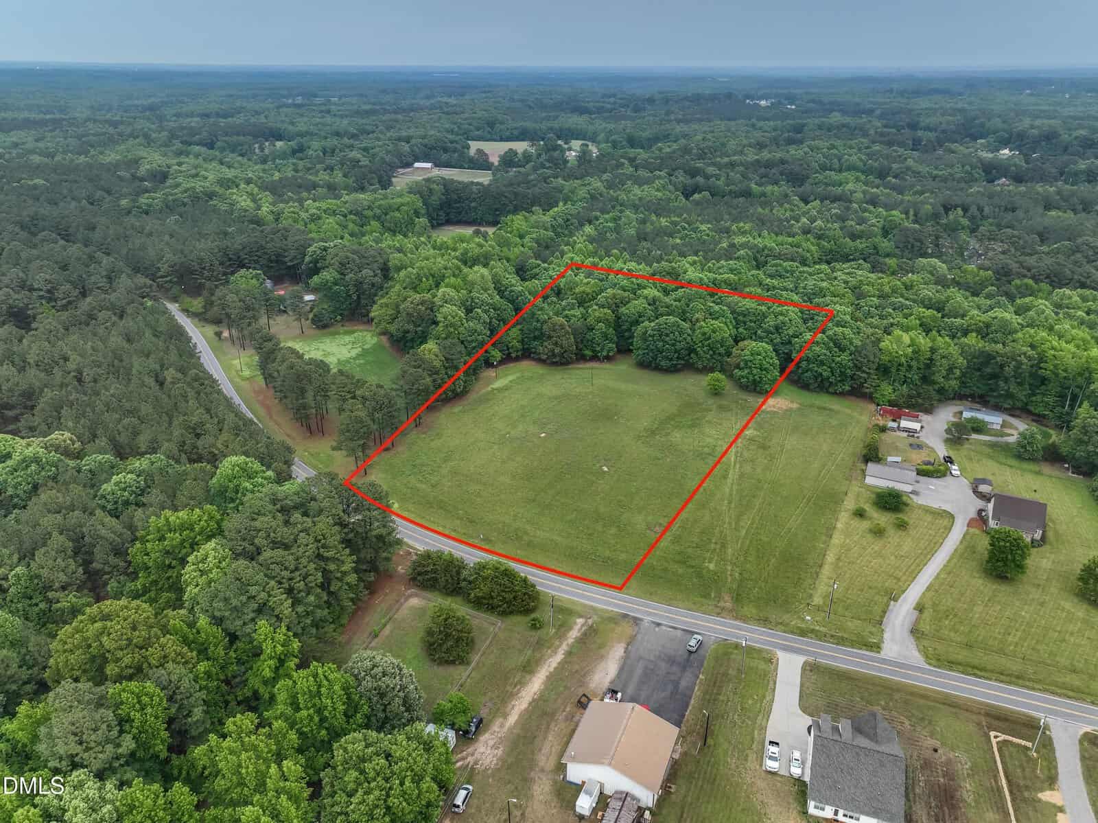 Aerial view of a grassy, rectangular plot of land outlined in red, surrounded by trees and near other homes, with roads bordering two sides, set in a rural, green landscape.