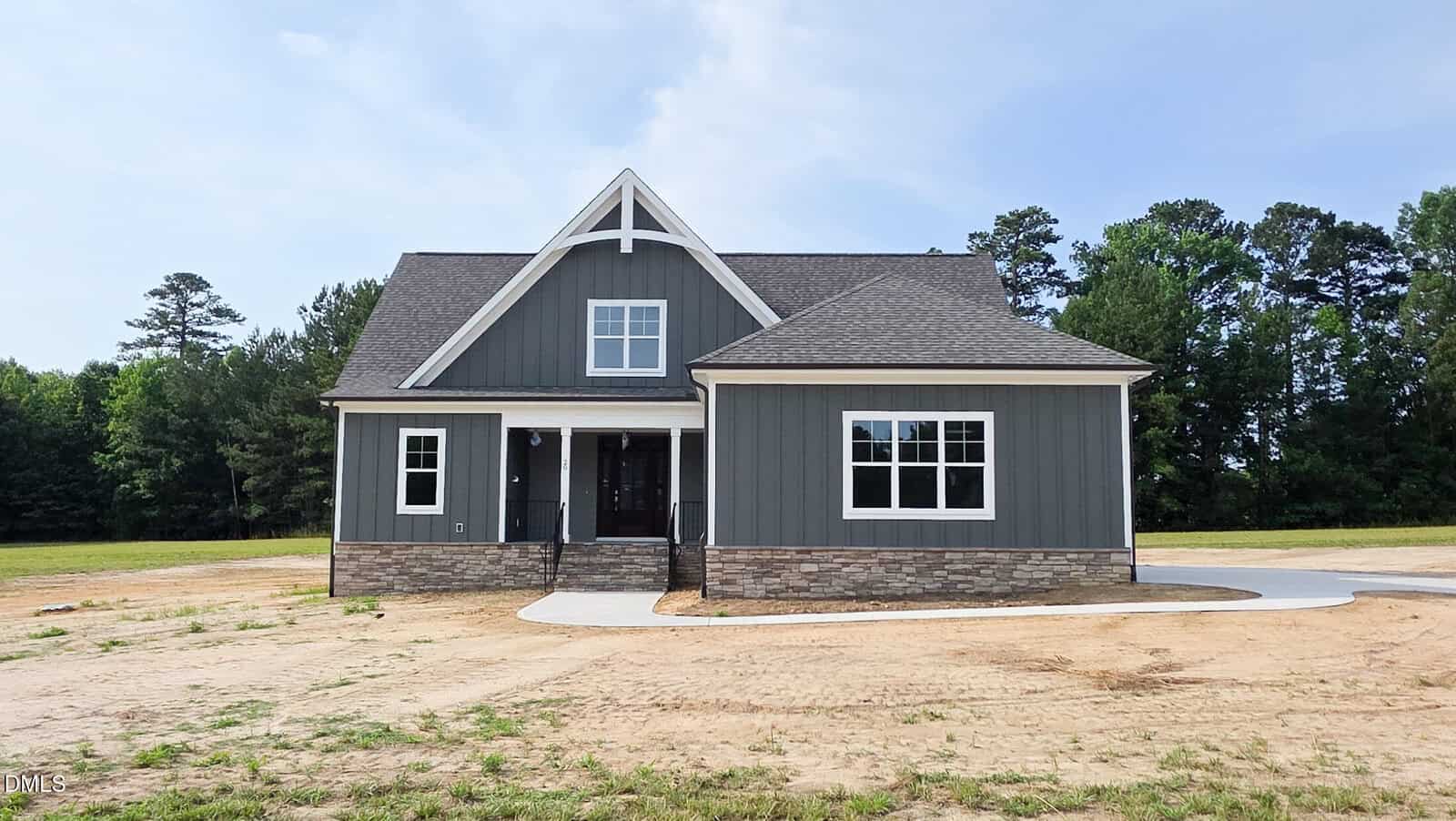 A modern gray house with white trim at 20 White Deer Lane, Youngsville, features a stone foundation and a peaked roof on a dirt lot with a partial concrete driveway, surrounded by trees under a blue sky.