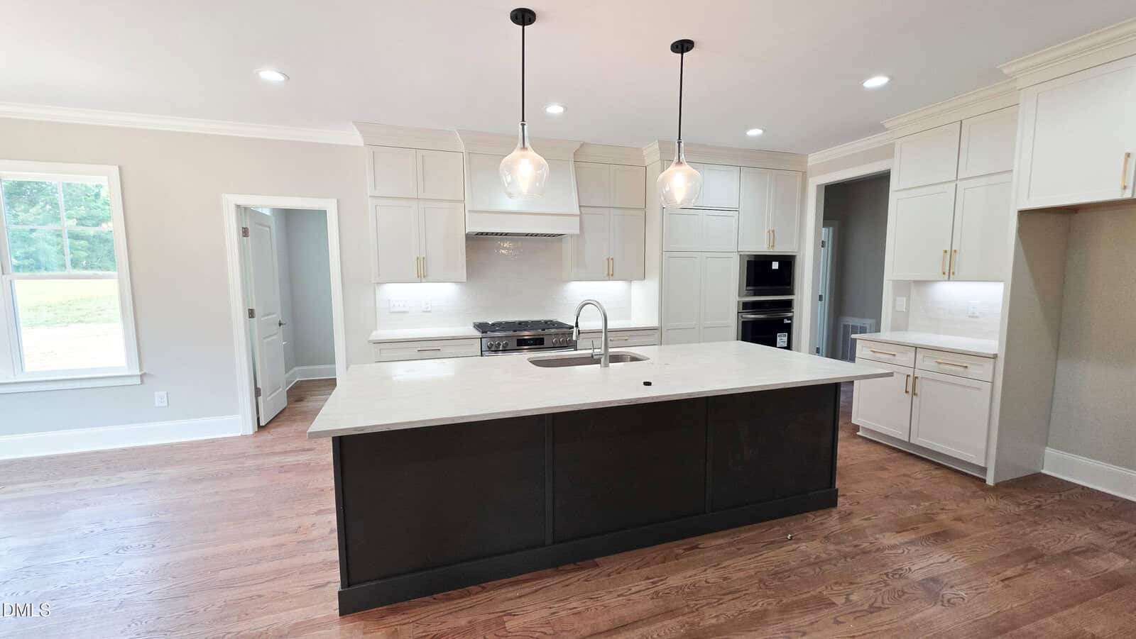 Modern kitchen with light cabinets, a dark island with a sink, pendant lights above, stainless steel appliances, and hardwood floors. A window brings in natural light from the left.