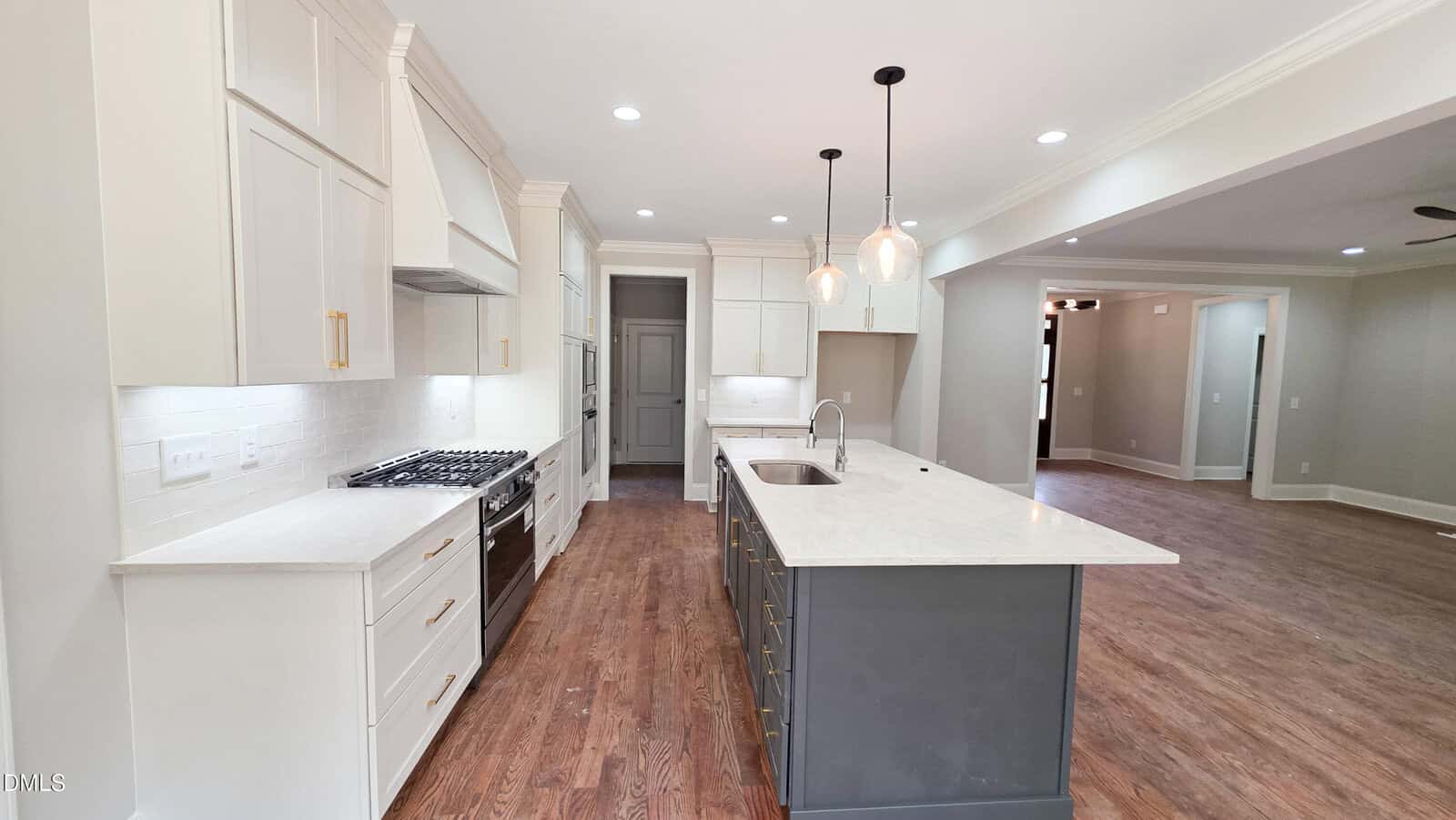 Modern kitchen with white cabinets, stainless steel appliances, a large gray island with sink, pendant lights, white countertops, and wood flooring. The kitchen opens into a spacious living area.