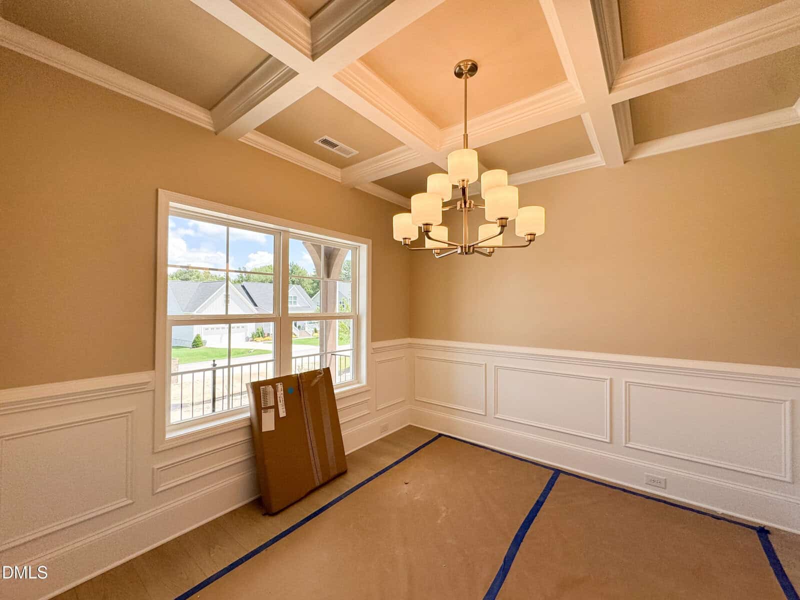 A dining room with beige walls, white wainscoting, a coffered ceiling, chandelier, and large window. The floor is covered with brown protective paper, and a large cardboard box leans against the wall.