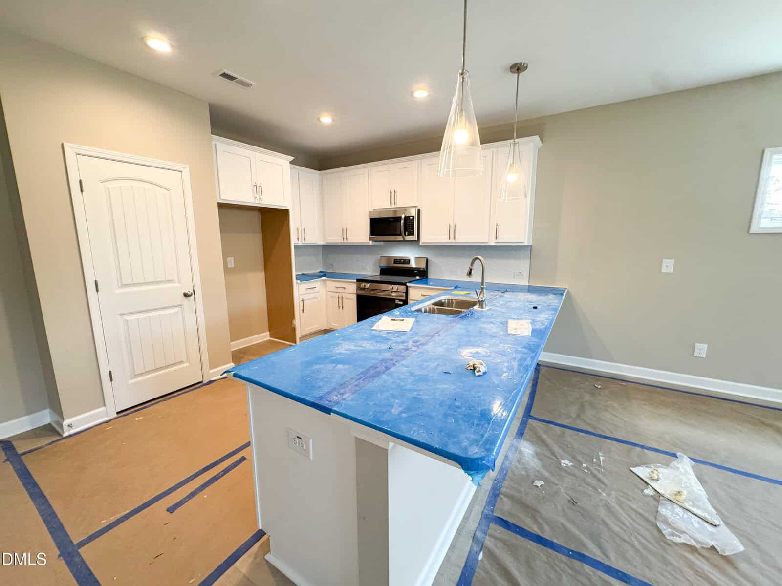 A modern kitchen under construction with white cabinets, stainless steel appliances, and a large island covered in blue protective wrap. Floors are covered with cardboard and tape.