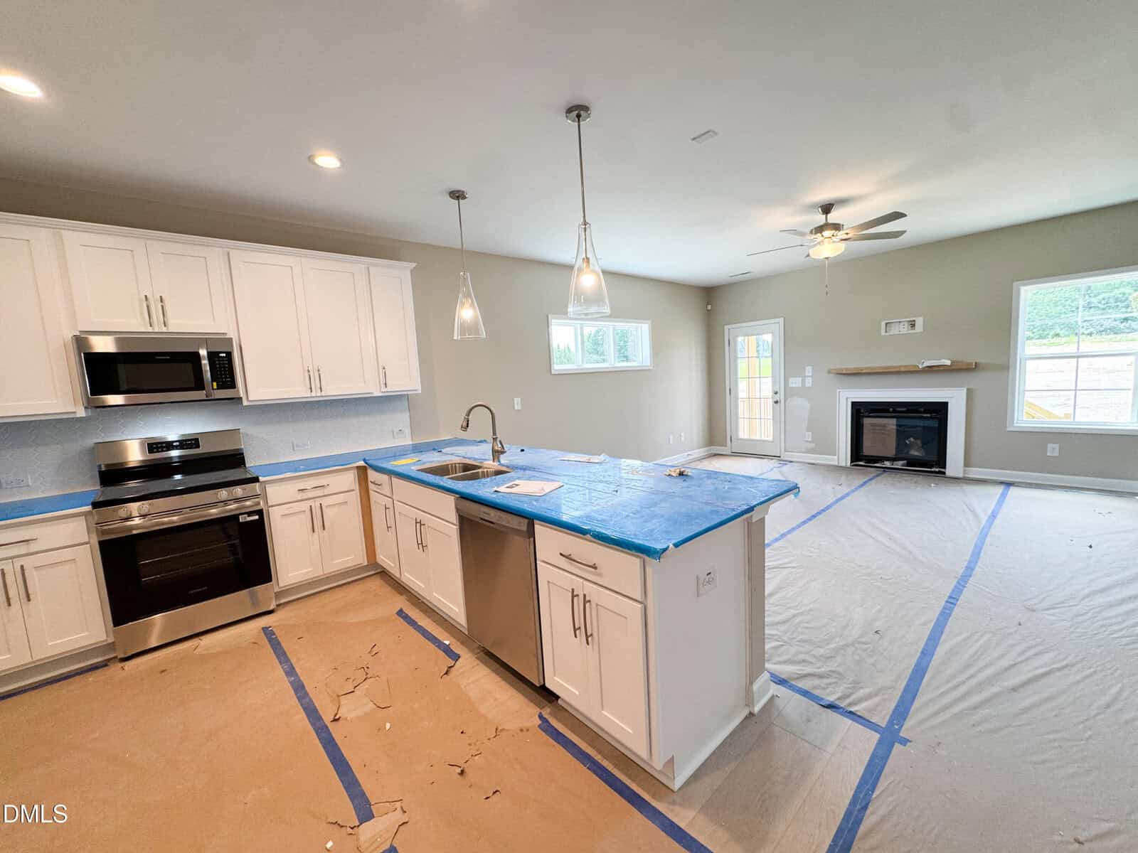 Newly constructed open-concept kitchen and living room with white cabinets, stainless steel appliances, and a covered floor for protection. Blue tape outlines kitchen island and living space. Ceiling lights and fan are visible.