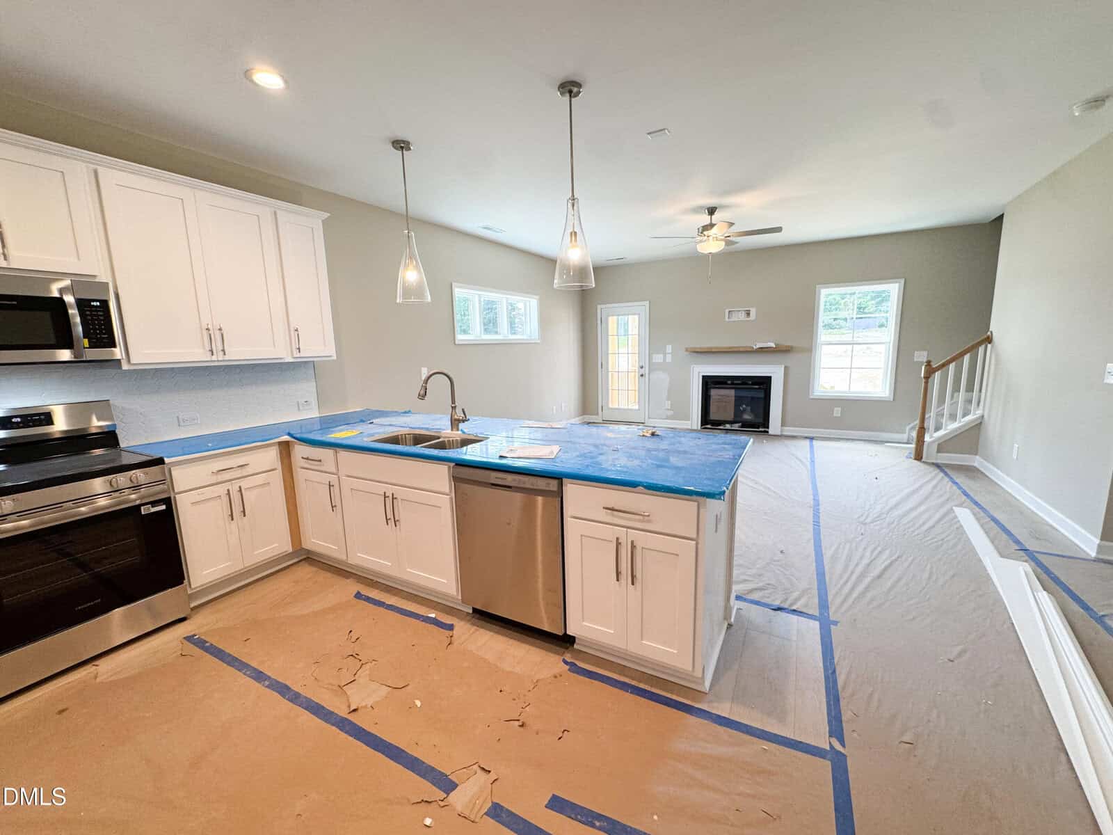A modern kitchen and living area under construction, with white cabinets, stainless steel appliances, blue coverings on counters, and protective paper on the floor leading to a fireplace and staircase.
