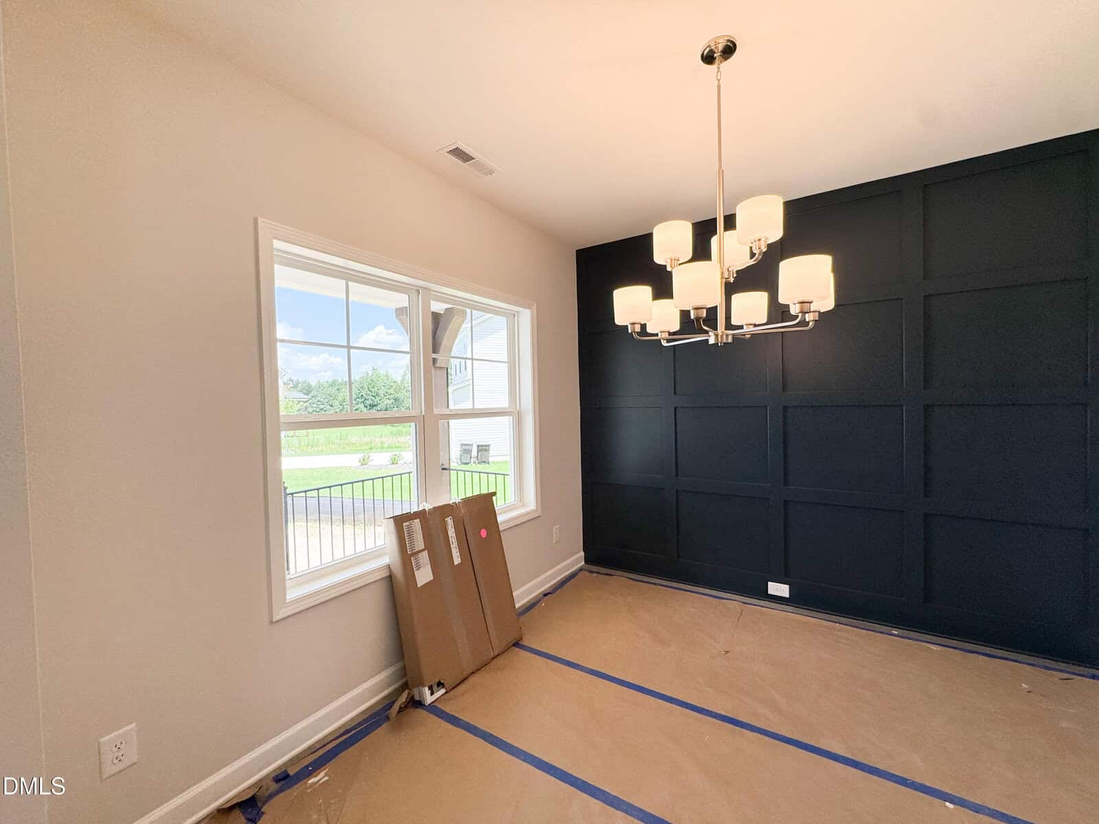 A bright room with a window, dark accent wall, modern chandelier, and cardboard-wrapped items on the floor. The floor is covered with protective paper, indicating the room is under renovation or recently constructed.