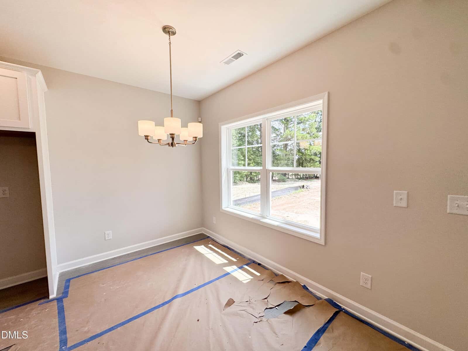 A bright, empty room with a large window, chandelier, and tan paper covering the floor. The walls are light beige, and there is blue tape along the floor edges, suggesting ongoing renovations.