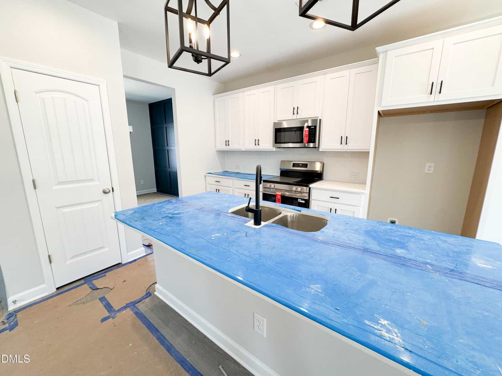 A modern kitchen with white cabinets, stainless steel appliances, and blue protective covering on countertops. A sink is installed on the island under black pendant lights. The floor is partially covered for protection.