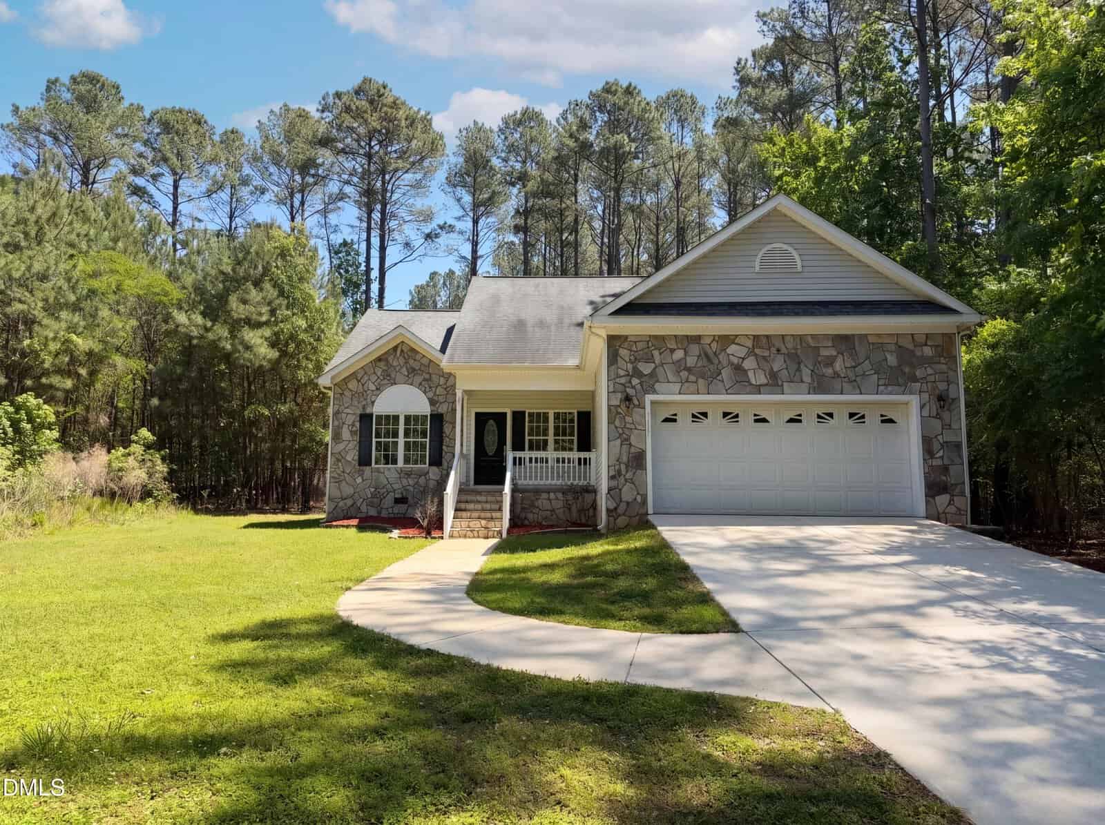 A single-story house with a stone facade, white trim, and a two-car garage sits beside a curved driveway, surrounded by green grass and tall trees under a partly cloudy sky.