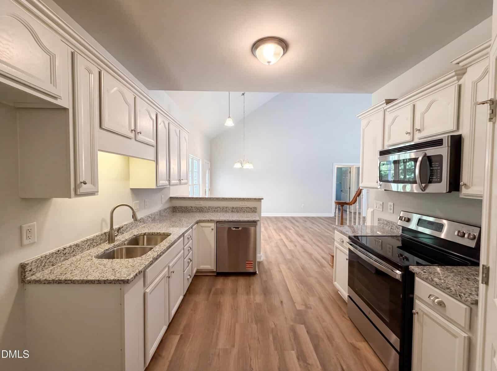 Modern kitchen with white cabinets, granite countertops, stainless steel appliances, a double sink, and wood-style flooring, leading to an open dining and living area with high vaulted ceilings and large windows.