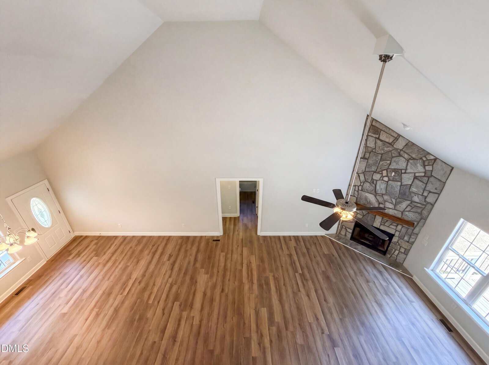 Spacious, empty living room with vaulted ceiling, wooden floors, a stone fireplace, large windows, ceiling fan, and a front door visible to the left. View is from above, capturing the open layout.