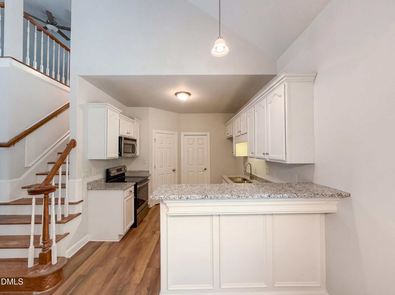 Bright kitchen with white cabinets, granite countertops, stainless steel appliances, wood flooring, and a breakfast bar. A staircase with wooden steps is visible on the left, and pendant lighting hangs from the ceiling.