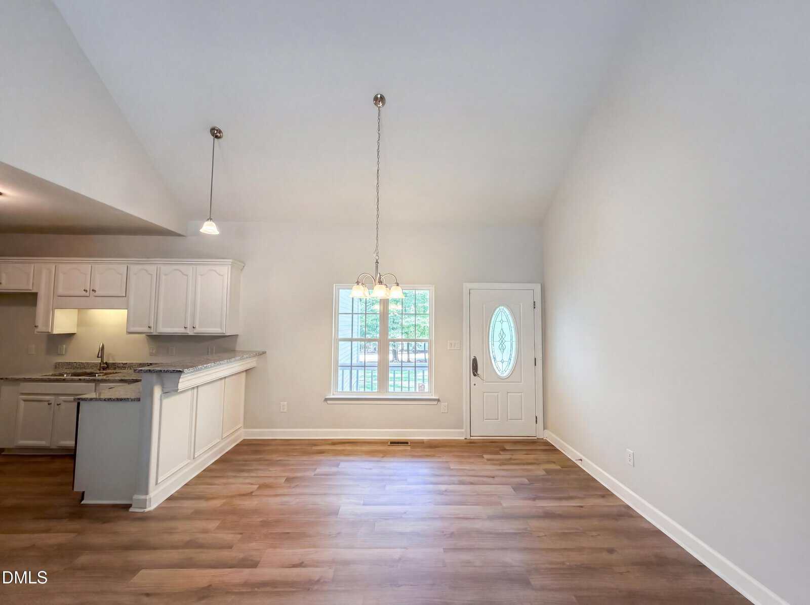 Open, bright living space with wood flooring, white cabinets, granite countertops, pendant lighting, a window, and a front door with an oval window. Light walls and high ceilings create an airy, spacious feel.