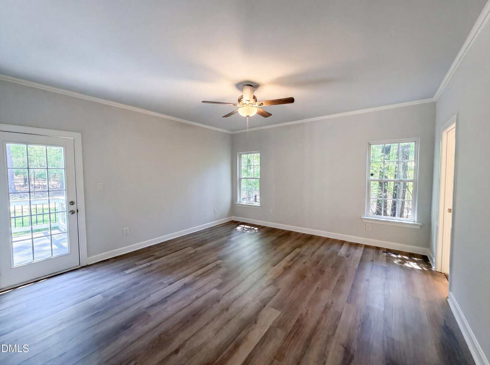 Bright, empty room with wood floors, light gray walls, a ceiling fan with lights, two windows, and a glass-paneled door leading outside. Natural light streams in from the windows and door.