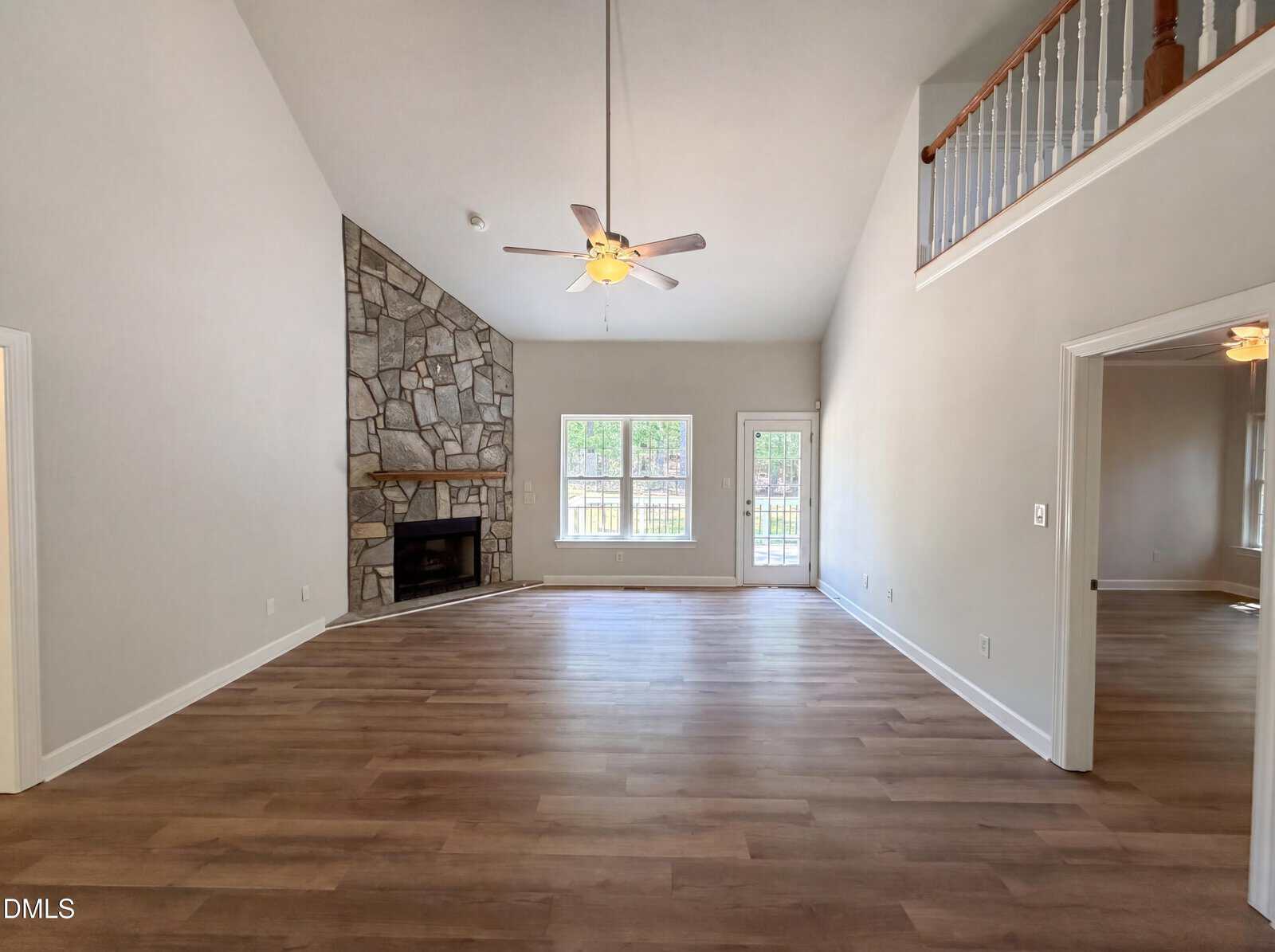 Spacious living room with light wood flooring, a stone fireplace in the corner, ceiling fan, large windows, and a door leading outside. An open walkway overlooks the room from the upper level.