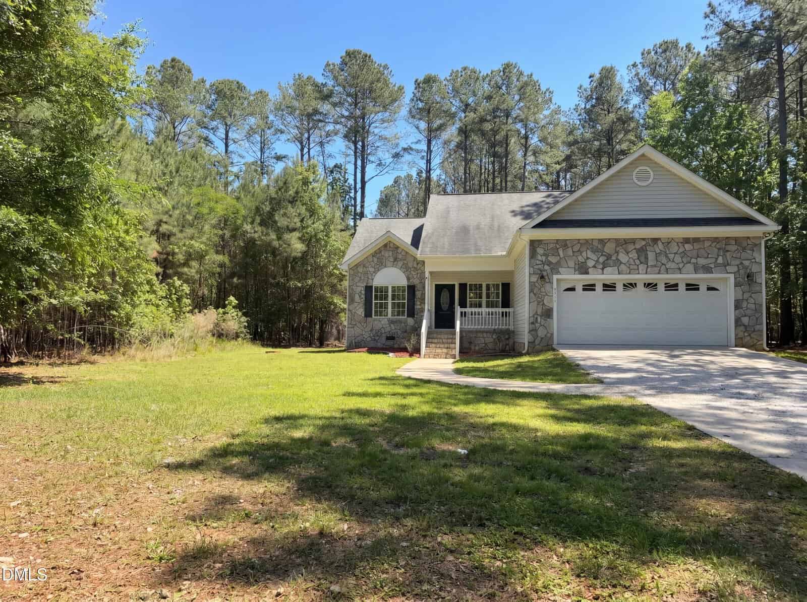 A single-story stone and siding house with a front porch and attached two-car garage sits on a grassy lawn, surrounded by tall trees under a clear blue sky.