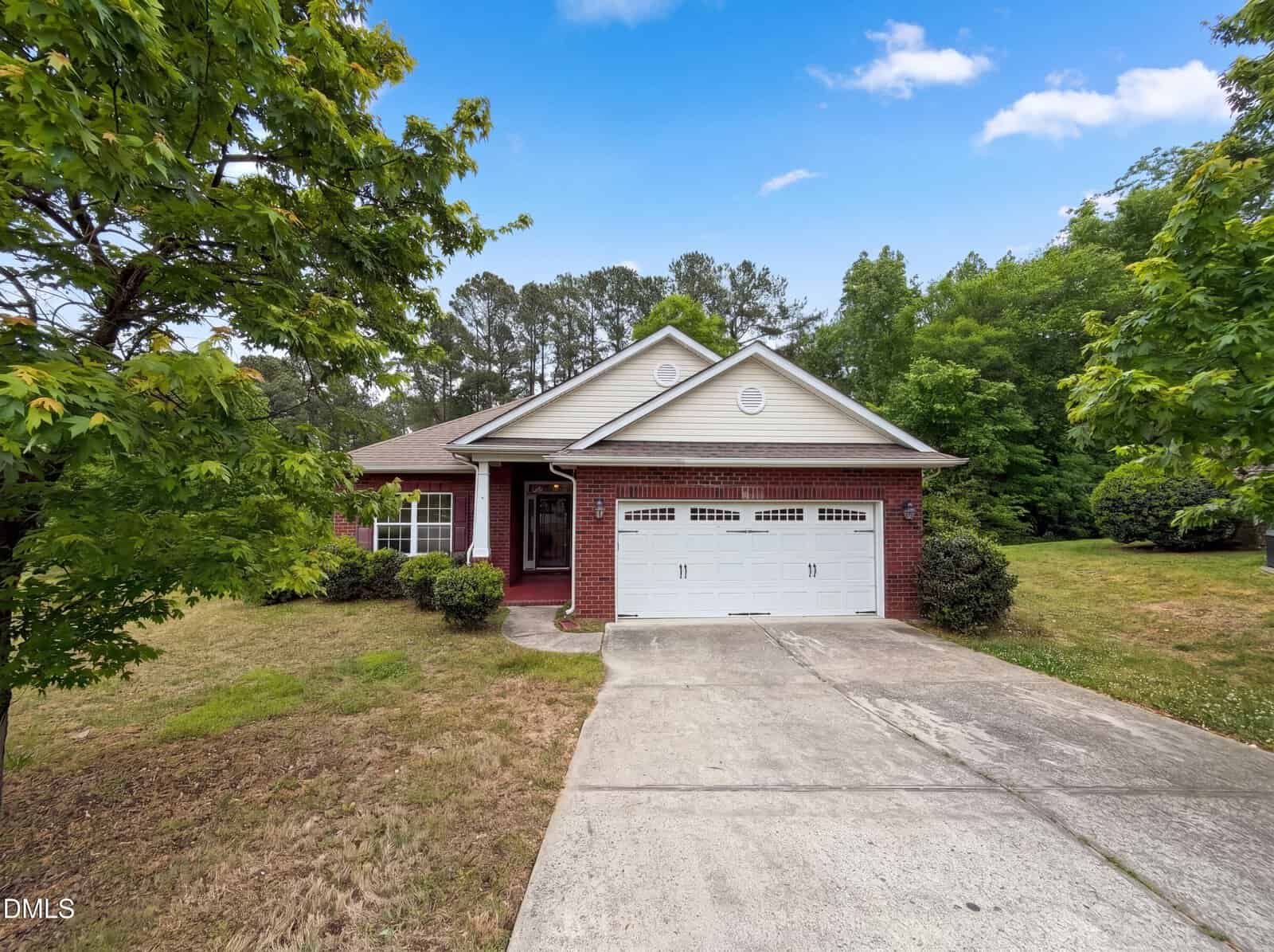 Single-story brick house with white trim, a double garage, and a driveway. Surrounded by green trees and grass, under a partly cloudy blue sky. Shrubs and plants line the front entryway.