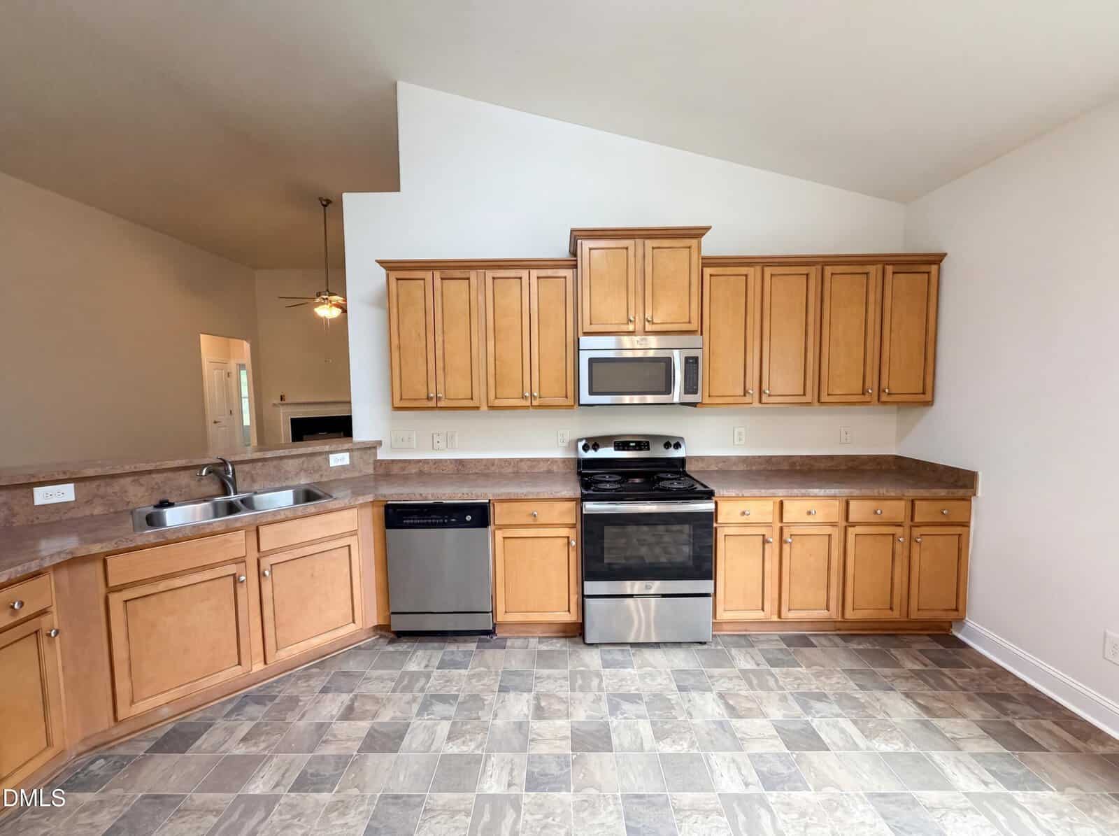 Spacious kitchen with light wood cabinets, stainless steel appliances including a stove, microwave, dishwasher, double sink, and neutral tile flooring. Walls are white and ceiling is vaulted.