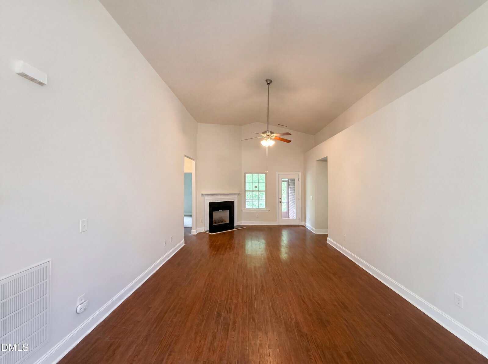 Spacious living room with high vaulted ceiling, ceiling fan with light, wood flooring, white walls, a corner fireplace, and windows letting in natural light at the far end.