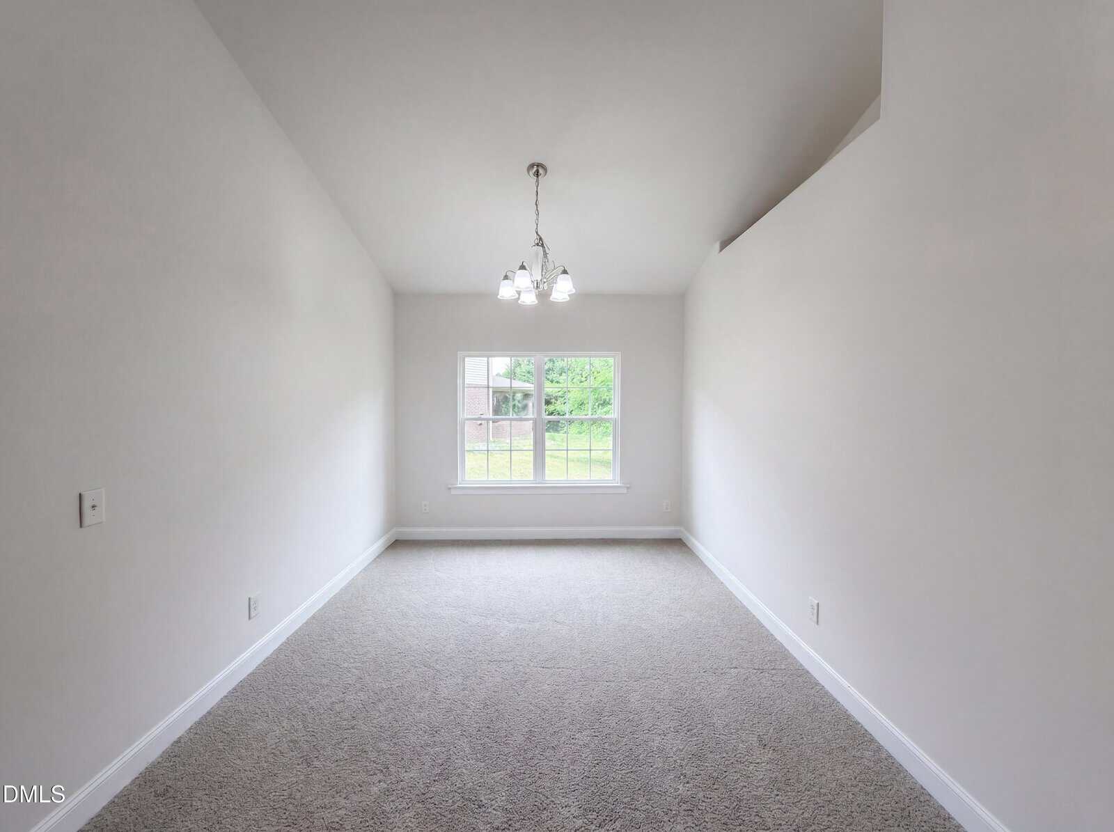 A bright, empty room with light gray carpet, white walls, a large window letting in natural light, and a modern chandelier hanging from the ceiling.