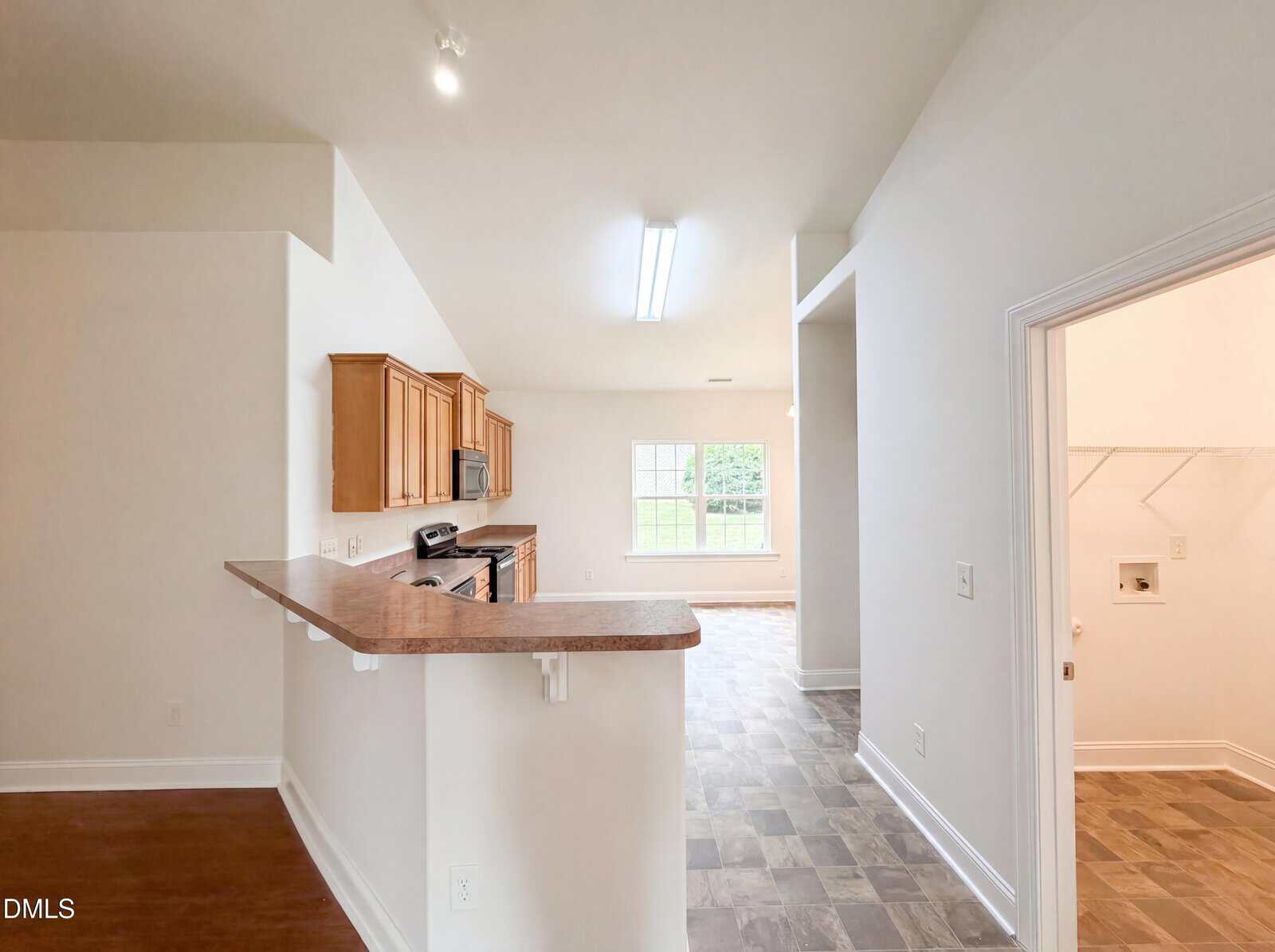 Open kitchen with wooden cabinets, brown countertops, and white walls; breakfast bar in foreground; adjacent laundry room with shelving; large window in dining area letting in natural light.