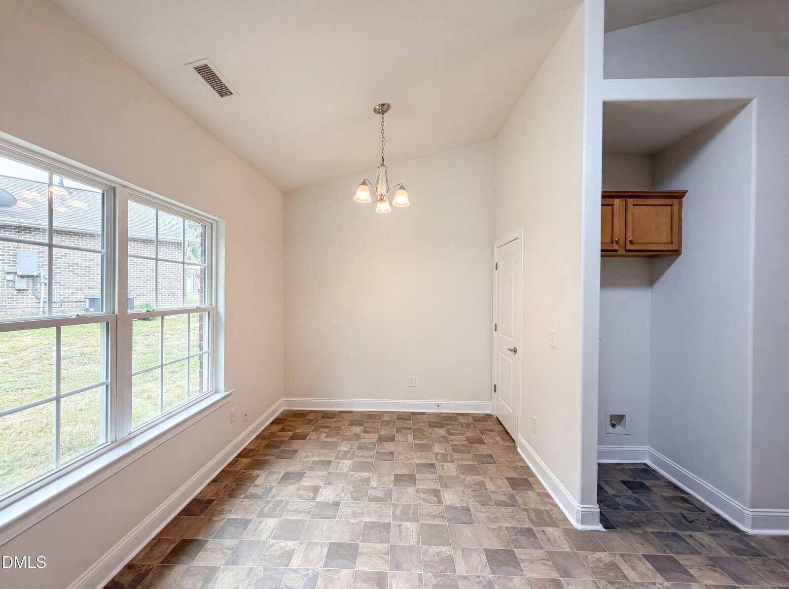 A bright dining area with a three-light chandelier, large window, light-colored walls, and tile flooring. There’s a small nook with a wooden cabinet and connections for a washer and dryer on the right.