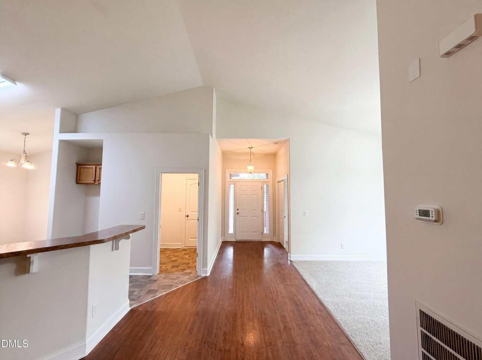 Spacious entryway of a house with vaulted ceilings, hardwood floors, and neutral-colored walls. The front door is at the center, with tile and carpeted areas on either side and a kitchen counter on the left.