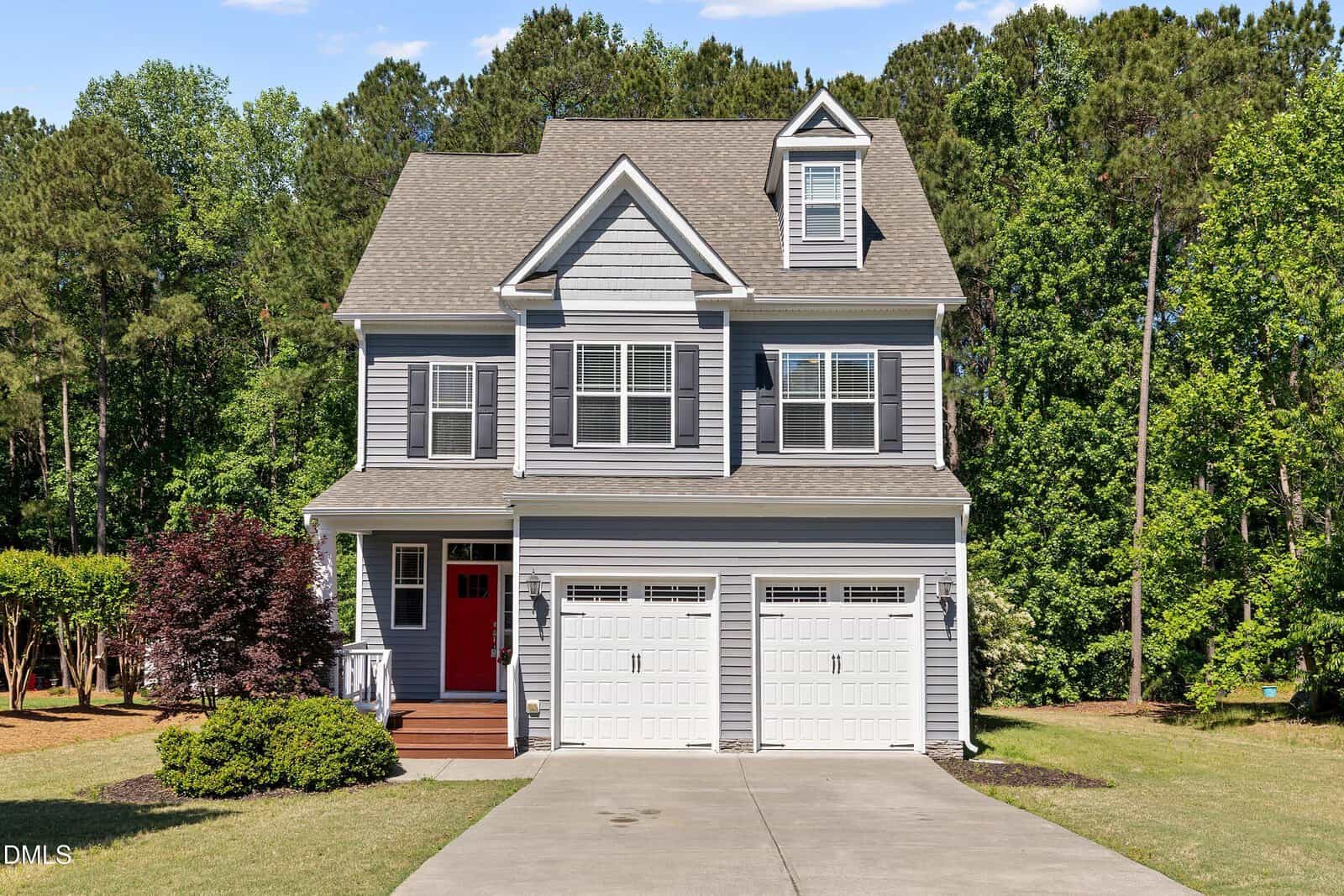 Two-story gray house with white trim, a red front door, and two white garage doors, surrounded by green trees and shrubs, with a concrete driveway leading up to the entrance.