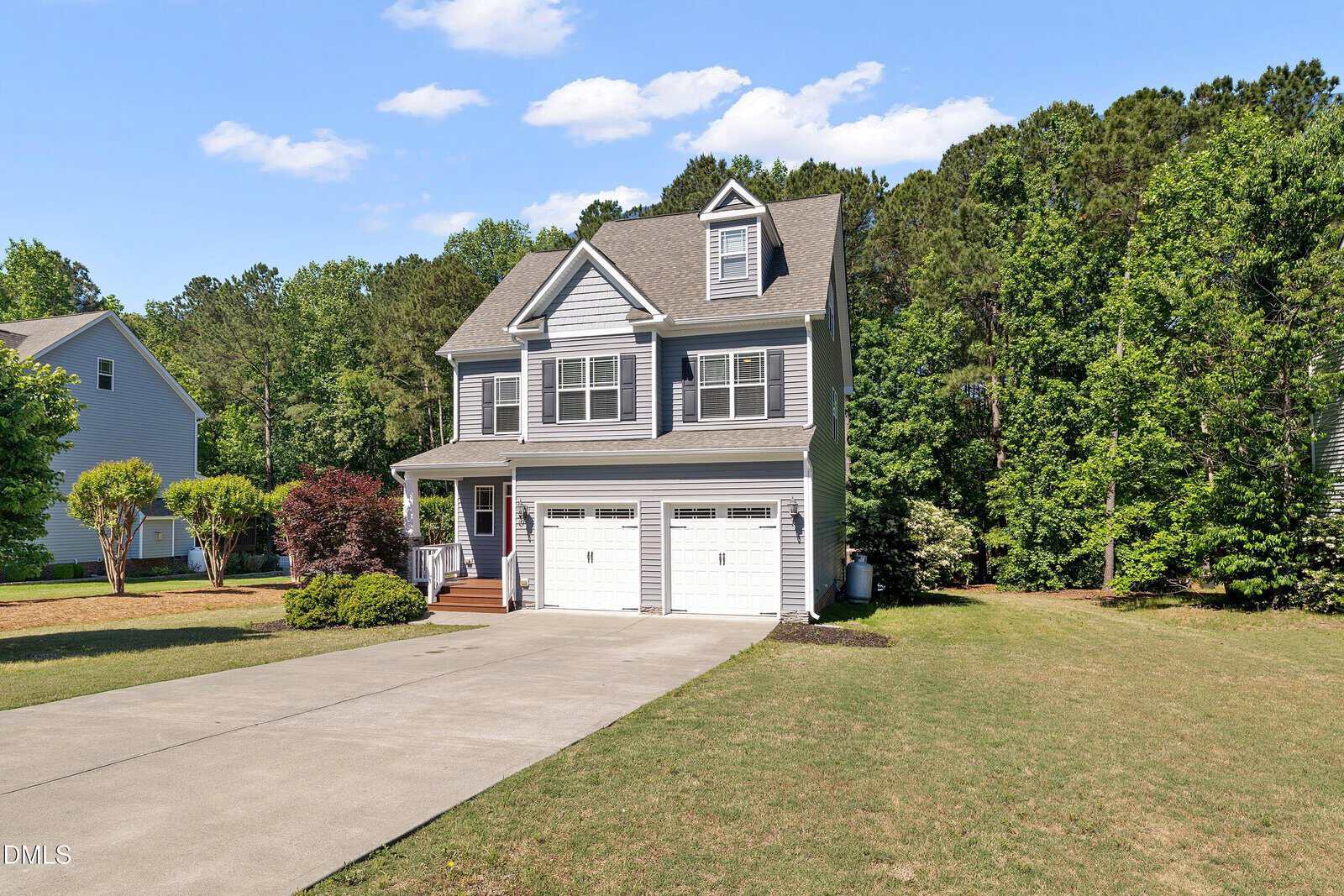 A two-story gray house with white trim, a covered front porch, and a double garage. The house is surrounded by green lawns, trees, and shrubs under a clear blue sky.