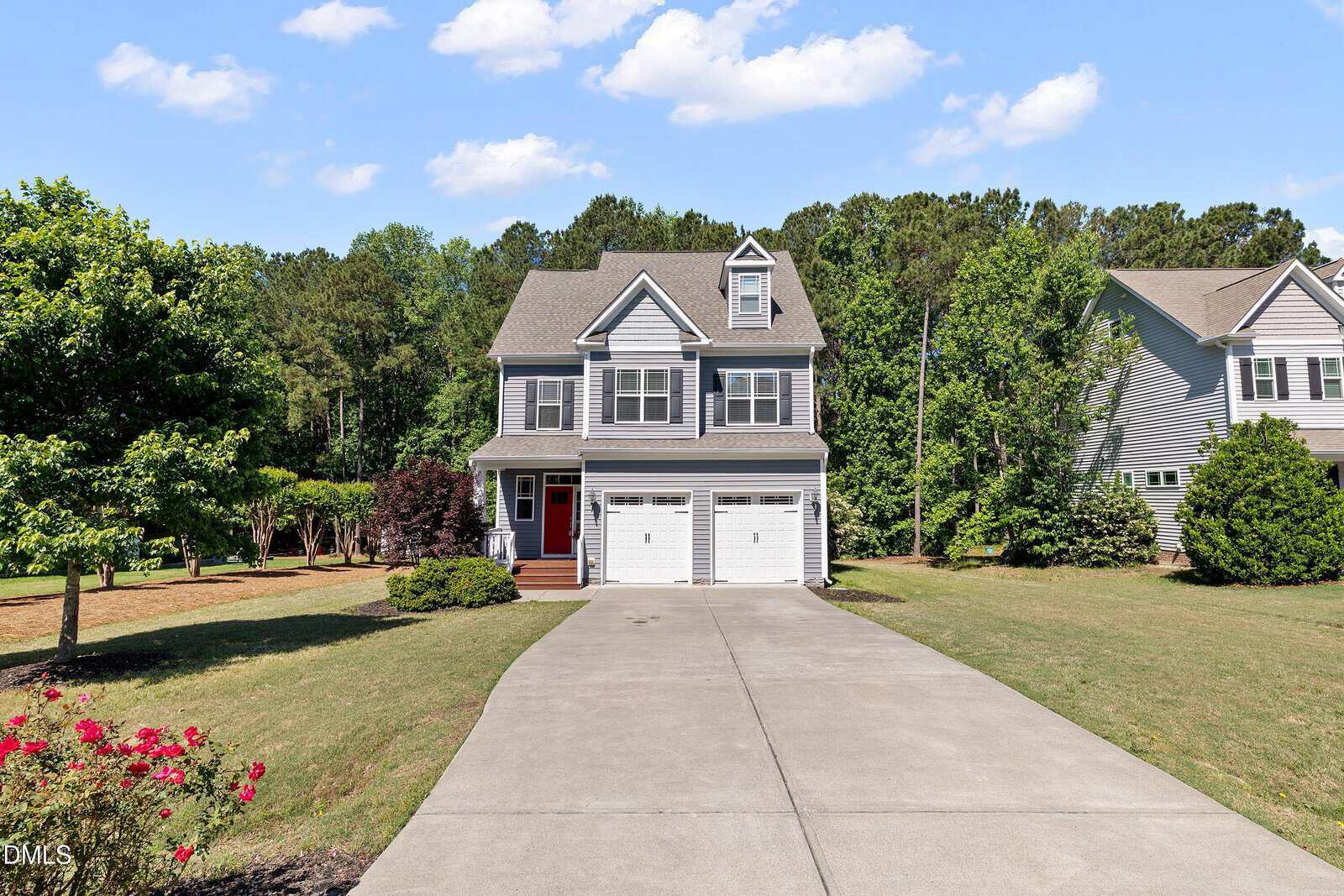 Two-story suburban house with gray siding, white trim, and a red front door, featuring a double garage and a wide driveway. The front yard is landscaped with grass, bushes, and trees, with a wooded area in the background.