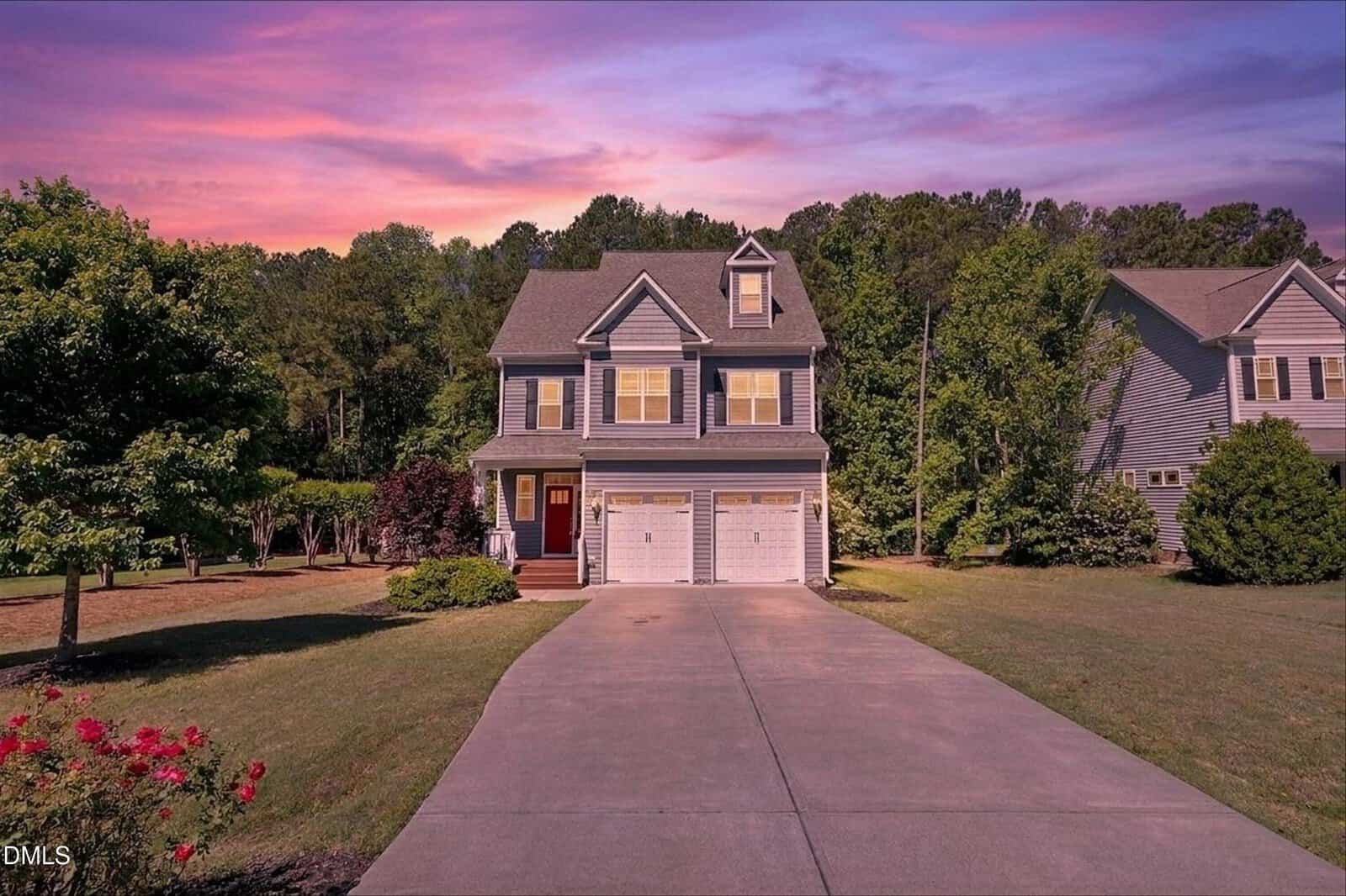 A two-story house with gray siding, white trim, and two garage doors sits at the end of a wide driveway, surrounded by a manicured lawn and trees, under a vibrant purple and pink sunset sky.