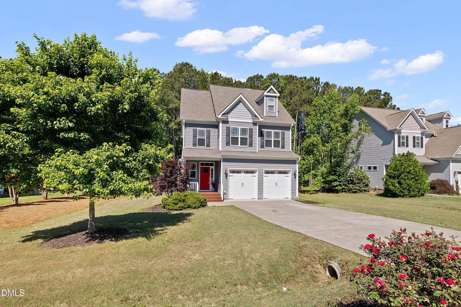 Two-story gray house with a red front door, attached double garage, and dormer windows. The home sits on a grassy lawn with trees and shrubs, under a blue sky with scattered clouds.